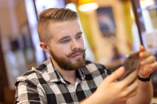 Стильный молодой человек с усами и бородой смотрит социальную сеть, используя смартфон, сидя в кафе на выходных. Концепция зависимости от социальных сетей . — стоковое фото