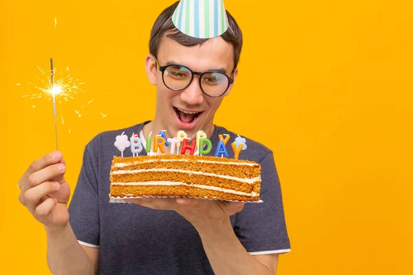 Сумасшедший веселый молодой азиат в очках с горящей свечой в руках и поздравительным домашним тортом на желтом фоне. Концепция празднования дня рождения и годовщины . — стоковое фото