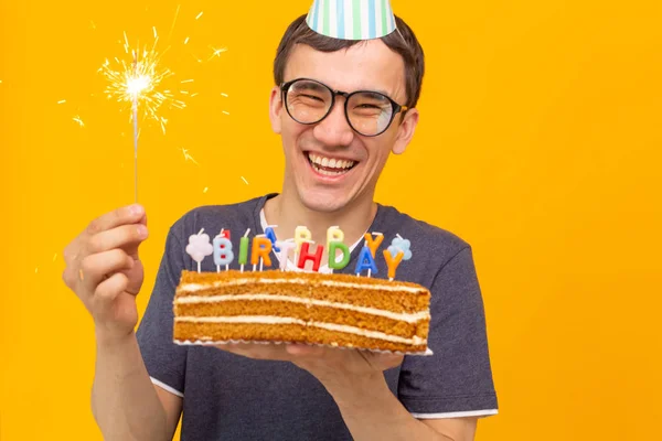 Fou joyeux jeune asiatique avec des lunettes tenant une bougie brûlante dans ses mains et un gâteau maison félicitant sur un fond jaune. Concept de célébration anniversaire et anniversaire . — Photo