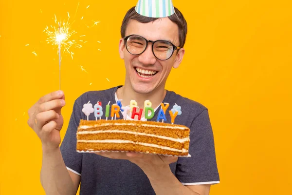 Pazzo allegro giovane ragazzo asiatico con gli occhiali che tiene una candela accesa tra le mani e una torta fatta in casa di congratulazioni su uno sfondo giallo. Concetto di compleanno e anniversario . — Foto Stock