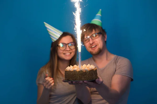 紙の帽子とケーキで面白い若いカップルは愚かな顔を作り、青い背景に立っている間、幸せな誕生日を願っています。おめでとうとだまさの概念. — ストック写真