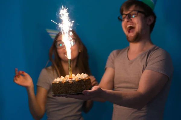 Joyeux jeune couple charmant mec et fille mignonne dans des chapeaux en papier faire visage stupide et tenir dans leurs mains un gâteau d'anniversaire debout sur un fond bleu . — Photo