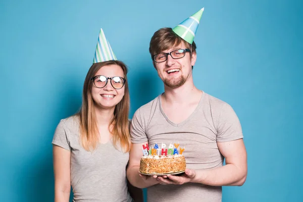 Funny Nerd mężczyzna i kobieta noszą wakacje czapki i okulary gospodarstwa tort urodzinowy ze świecami na niebieskim tle — Zdjęcie stockowe