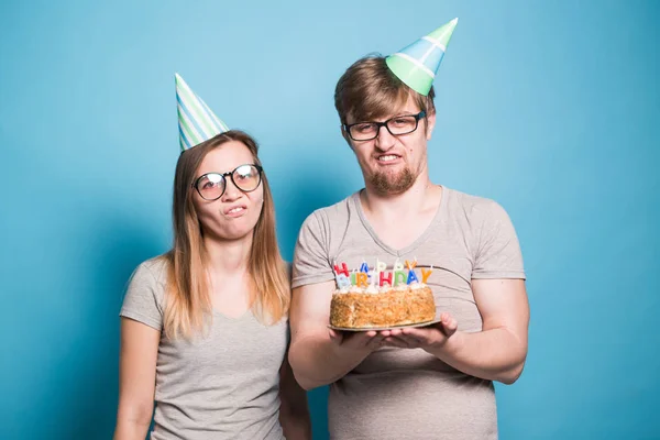 Alegre joven pareja encantador chico y lindo chica en papel sombreros hacer tonto cara y mantener en sus manos un pastel de cumpleaños de pie sobre un fondo azul . —  Fotos de Stock