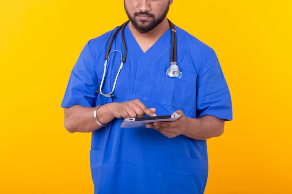 청진기를 가진 파란색 가운을 입은 귀여운 인도 의사가 노란색 배경을 기반으로 태블릿을 보고 있습니다. 컨셉 온라인 상담 — 스톡 사진