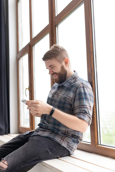 Cute młody facet Hipster ze stylowymi ubraniami siedzi na sieciach społecznościowych za pomocą smartfona i bezprzewodowego Internetu w pobliżu dużych okien. Koncepcja uzależnienia od Internetu. — Zdjęcie stockowe