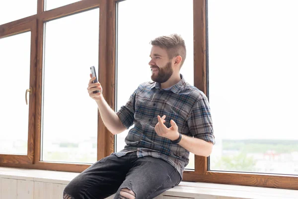 Lindo chico positivo joven hipster con una barba está sentado en el alféizar de la ventana grande y hablando en la comunicación de vídeo utilizando un teléfono inteligente e Internet inalámbrico de alta velocidad . — Foto de Stock