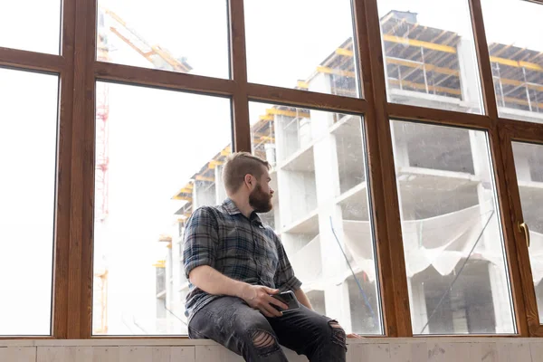 Joven hombre guapo hipster sentado en el alféizar de la ventana cerca de una gran ventana con un teléfono inteligente en sus manos y mirando el sitio de construcción en casa. Concepto de arquitecto novato . — Foto de Stock