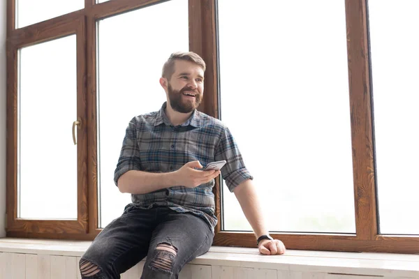 Νέος όμορφος άντρας χίπης κάθεται στο περβάζι κοντά σε ένα μεγάλο παράθυρο με ένα smartphone στα χέρια του και κοιτάζοντας το εργοτάξιο στο σπίτι. Ιδέα του πρωτάρη αρχιτέκτονα. — Φωτογραφία Αρχείου