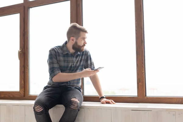Νέος όμορφος άντρας χίπης κάθεται στο περβάζι κοντά σε ένα μεγάλο παράθυρο με ένα smartphone στα χέρια του και κοιτάζοντας το εργοτάξιο στο σπίτι. Ιδέα του πρωτάρη αρχιτέκτονα. — Φωτογραφία Αρχείου