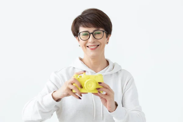 戴着眼镜的中年妇女手里拿着一个老式的黄色相机,在白色背景上摆姿势。摄影爱好者的概念和爱好 — 图库照片