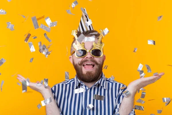 Joven hipster positivo loco con barba se ríe alegremente entre los kofetti voladores sobre un fondo amarillo. El concepto de vacaciones y ventas — Foto de Stock