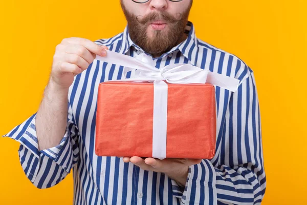 Surprised young hipster man with mustache and beard in surprise unpacks a red box with gifts on a yellow background, close up. The concept of gifts and surprises for the holiday