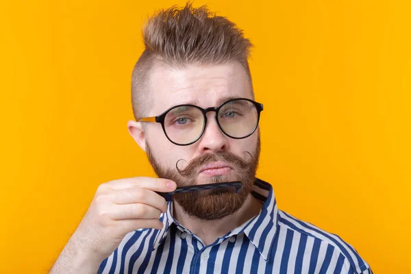 Retrato de un joven y guapo hipster con bigote y barba peinándose el bigote sobre un fondo amarillo. El concepto de barbería y belleza . — Foto de Stock