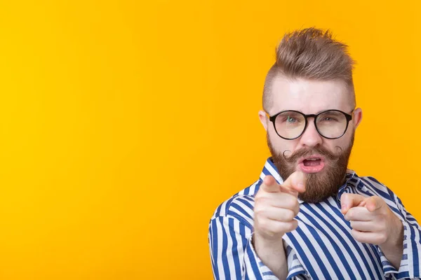 Funny young man with a mustache and a beard and glasses shows on you on a yellow background with copy space. Concept of non-standard advertising.