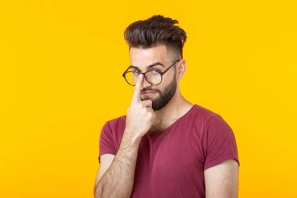 Homem hipster jovem e pensativo com uma barba segurando seus óculos enquanto estava de pé contra um fundo amarelo. O conceito de atenção e escolha difícil — Fotografia de Stock