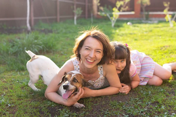 Mignon jeune mère et fille s'asseoir sur l'herbe de leur maison de campagne pieds nus à côté de leur chien bien-aimé par une journée d'été ensoleillée. Concept de vacances familiales . — Photo