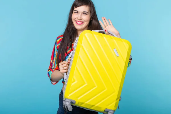 Portrait d'une belle jeune femme brune avec une valise jaune dans les mains posant sur un fond bleu. Concept de voyage et de tourisme . — Photo