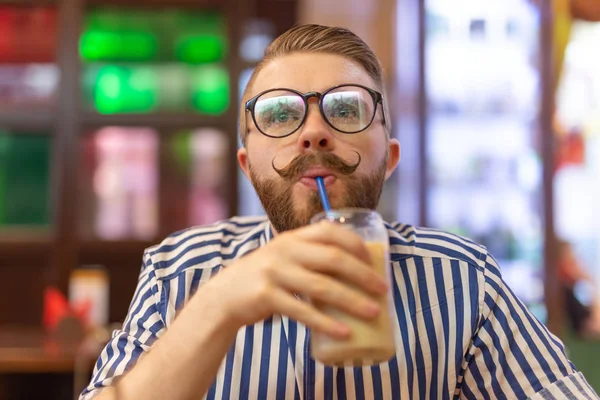 Retrato de un joven guapo con bigote y barba bebiendo café helado en un café sobre un fondo borroso. El concepto de estudiante o los hombres con estilo . — Foto de Stock
