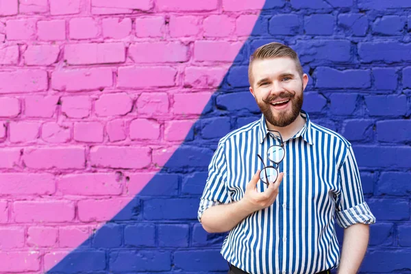 Retrato de joven alegre positivo elegante con bigote y barba y gafas en las manos posando sobre fondo de pared de ladrillo azul-violeta con espacio de copia — Foto de Stock