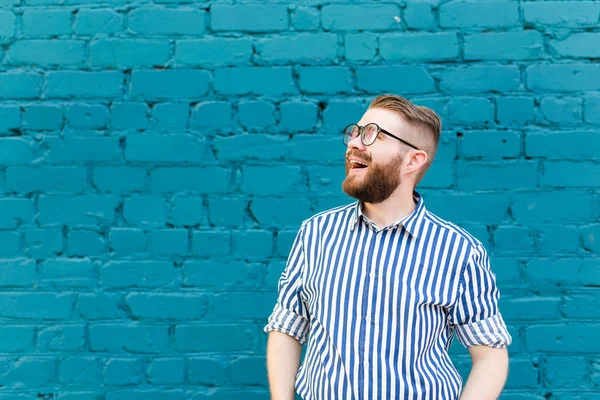 Portret van een jonge positieve stijlvolle lachende kerel met een snor en baard tegen de achtergrond van een blauwe bakstenen muur met Kopieer ruimte. Vakantie concept in de stad. — Stockfoto