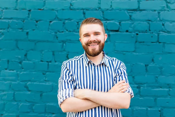 Retrato de un joven chico sonriente positivo con un bigote y barba contra el fondo de una pared de ladrillo azul. Concepto de vacaciones en la ciudad . — Foto de Stock