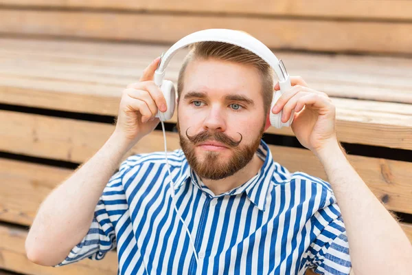 Portret van een knappe jonge studente jongen met een snor en een baard ontspannen in een park met een koptelefoon op een warme zomerdag. Weekend uitje concept. — Stockfoto