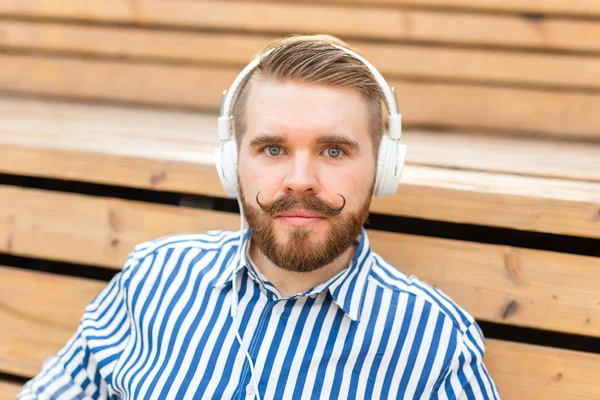 Portret van een knappe jonge studente jongen met een snor en een baard ontspannen in een park met een koptelefoon op een warme zomerdag. Weekend uitje concept. — Stockfoto