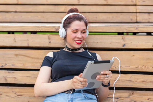 Portrait of a beautiful stylish young cheerful woman in headphones sitting in the park on a warm summer day. The concept of leisure and study at the weekend. — Stock Photo, Image