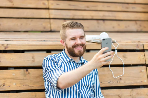 Positieve jonge kerel student met snor en baard praten met zijn vrienden op video communicatie met behulp van smartphone en gratis Wi-Fi internet zittend in het Park op een bankje met een Tablet en een koptelefoon. — Stockfoto