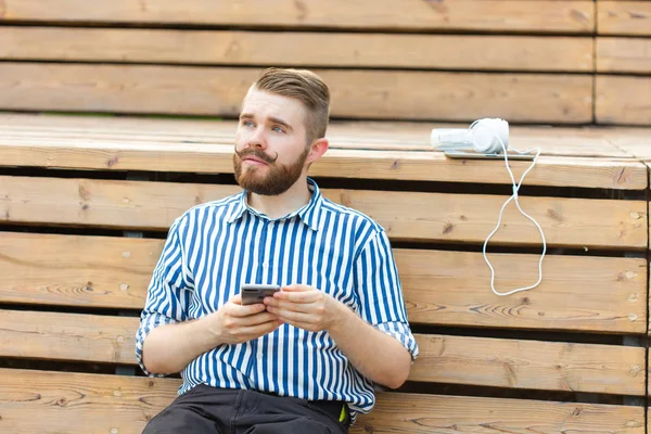 Pensativo joven estudiante hipster masculino serio con un bigote y barba escribiendo sms mensaje a sus amigos descansando después de estudiar en el parque sentado en un banco de madera con una tableta y auriculares — Foto de Stock