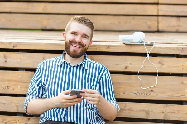 Pensativo joven estudiante hipster masculino serio con un bigote y barba escribiendo sms mensaje a sus amigos descansando después de estudiar en el parque sentado en un banco de madera con una tableta y auriculares — Foto de Stock