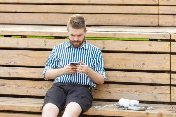Pensive poważne młody mężczyzna student Hipster z wąsy i brody piśmie wiadomości SMS do swoich przyjaciół odpoczynku po studiach w parku siedzi na drewnianej ławce z tabletem i słuchawkami — Zdjęcie stockowe