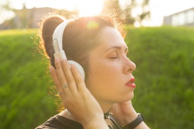 Genç güzel kadın sıcak bir yaz akşamı parkta yürürken kulaklık ve online radyo kullanarak rahatlatıcı müzik dinlerken. Müzik abonelik konsepti.
