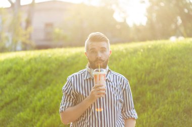 Gözlük ve şık sakallı pozitif genç hipster adam bir parkın karşısında dışarıda dururken bir saman ile milkshake içiyor. Hafif tatlı içecekler sevenler kavramı.