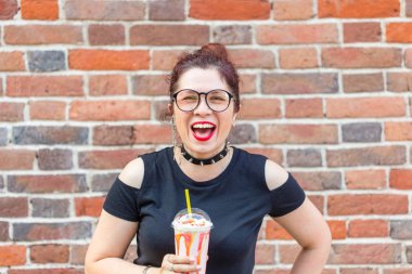 Büyüleyici neşeli genç şık kız bir tuğla duvar arka plan üzerinde bir saman ile milkshake tutan. Bir kafede ve sokakta yaz tatili kavramı.