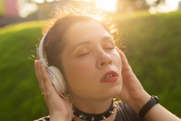 Beautiful sensual young woman listens to music with headphones while walking through the park on a sunny evening. Weekend getaway concept.
