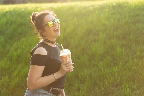 Pozytywna, wesoła młoda kobieta w stylowych punkowych ubraniach i okularach trzyma koktajl w rękach podczas spaceru w parku w ciepłym letnim wieczorem. Koncepcja relaksu i zabawy w mieście. — Zdjęcie stockowe