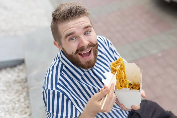 Zblízka na zábavného mladého hipstera, který jedl čínské nudle s dřevěnými hůlky sedících v parku venku za teplého letního dne. Koncept odpočinku a zachvívání na ulici. — Stock fotografie