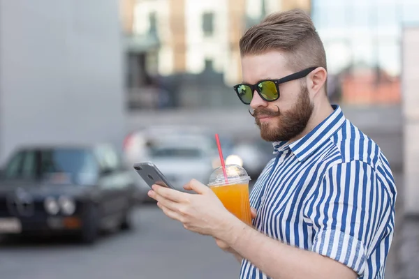 Widok z boku młodego stylowego Hipster człowiek z okularami i wąsy i sok w ręce rozmowy w sieciach społecznościowych za pomocą smartfona i bezprzewodowego Internetu w ciepłym letnim dniu. — Zdjęcie stockowe