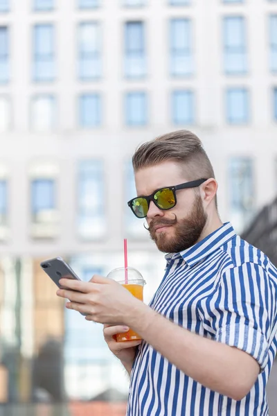 Vista lateral de un joven y elegante hombre hipster con gafas y un bigote y jugo en sus manos charlando en las redes sociales usando un teléfono inteligente e internet inalámbrico en un cálido día de verano . — Foto de Stock