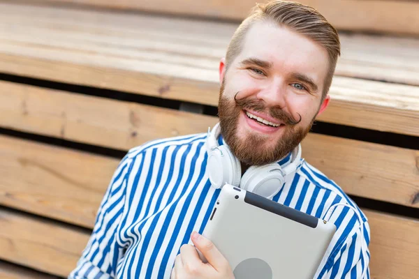 Retrato de un joven estudiante guapo con bigote y barba relajándose en un parque con auriculares en un cálido día de verano. Concepto de escapada de fin de semana . — Foto de Stock