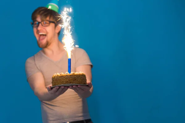 Ein verrückter, fröhlicher Kerl mit Brille grüßt mit einem Papierhut, der eine Torte mit brennenden Feuerwerkskörpern in den Händen hält. Feriengeburtstag-Konzept — Stockfoto