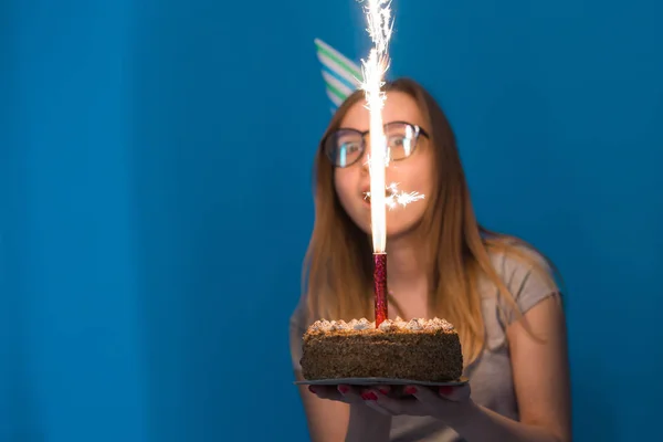 青い背景にキャンドルを持つお祝いのケーキを持って眼鏡をかけた陽気な若い女子学生。誕生日のコンセプト. — ストック写真