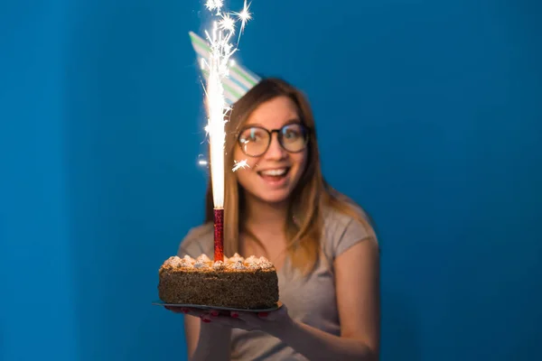 Vidám ifjú homályos lány hallgató szemüveg kezében gratuláló torta egy gyertyát állva a kék háttérben. Születésnapi koncepció. — Stock Fotó