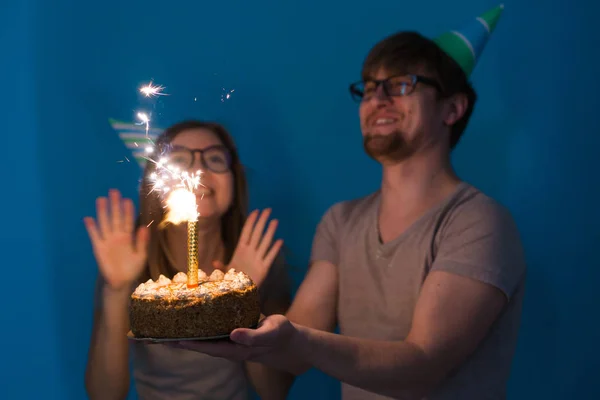 Hombre y mujer nerd divertido están usando gorras y gafas de vacaciones celebración de pastel de cumpleaños con espumoso sobre fondo azul —  Fotos de Stock