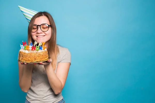 Uroczy wesoły szalony młoda dziewczyna uczeń w gratulacyjny kapelusz papieru gospodarstwa szczęśliwy tort urodzinowy w jej ręce stoi na niebieskim tle. Przestrzeń reklamowa. — Zdjęcie stockowe