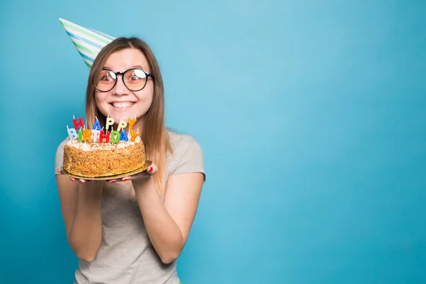 Закройте смешную позитивную девушку в очках и приветственной бумажной шляпе, держа в руках праздничный торт, стоящий на синем фоне с копировальным местом — стоковое фото