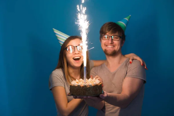 Смішний ботанік і жінка носять шапочки і окуляри, що тримають торт на день народження зі свічками на синьому фоні — стокове фото