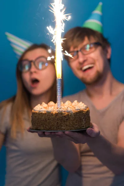 Divertido nerd hombre y mujer están usando días festivos gorras y gafas celebración de pastel de cumpleaños con velas sobre fondo azul —  Fotos de Stock